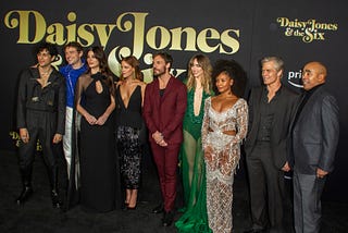 The cast of Daisy Jones and the Six on a black carpet in front of a black and gold background.