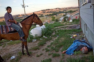 Photo: Aaron Huey → young boy on horseback looks down and local reservation member passed out behind the house from over drinking.