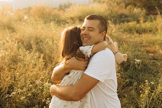A father hugs his school-aged daughter, smiles, and closes his eyes. The daughter holds a stuffed animal in one hand. The photo is set in a field at sunset, and the plants in the background are illuminated by the setting sun.