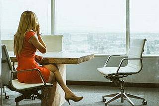 A woman, back to camera, sits at a conference table while talking on a cell phone and looking out of a highrise at a cityscape.