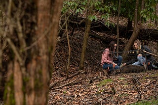 Two women bending over in a forest inspecting a dead pig in a cage.