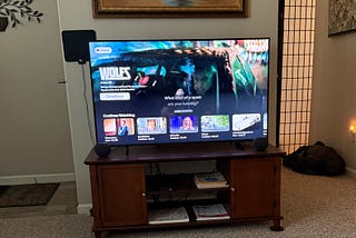 A living room setup with a large flat-screen TV on a wooden stand displaying a movie selection screen. The TV shows the movie Wolfs with the tagline, “What kind of a scam are you running?” Below the main selection, there are smaller options for movies and TV shows like Elementary, Bad Sisters, Lie to Me, and Nature. The TV stand has a dark wood finish with shelves below holding books and an Apple TV box. Small black speakers are positioned on either side.