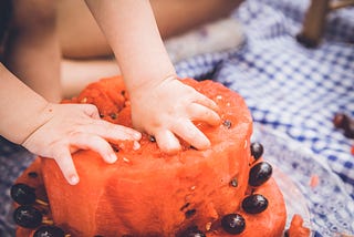 A toddler stocks their hands into a fresh fruit cake made with watermelon and grapes.