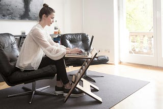 Woman wearing white flowy top and black pants typing on laptop while sitting on black leather chair in living room.