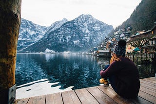 A woman sits on a deck and looks at a lake in winter. She is wearing warm clothes. A mountain in the distance is covered with snow. | Photo courtesy: Riccardo, Pexels