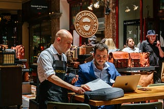 Small-business owner reviews documents with a retail associate while working on a laptop in a shop setting. Photo by Johan Mouchet on Unsplash