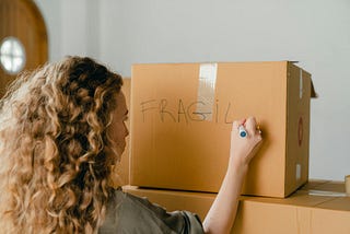 A woman with curly strawberry blonde hair writes “fragile” with a marker on a cardboard moving box.