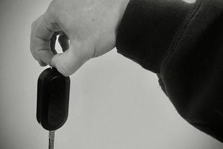 A black and white photo of an arm outstretched, dangling  a car key in their fingers.