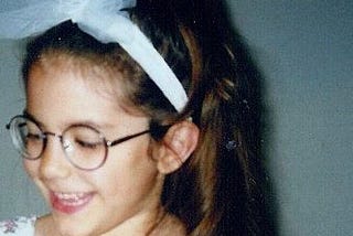 A photo of the author at age four, wearing glasses, a long brunette ponytail, and a white tulle headband.