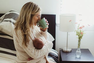 Mid adult woman sitting on bed drinking coffee whilst cradling new born baby daughter