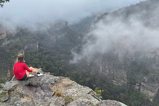 Woman with shoulder length brown hair wearing red top, black shorts and white sneakers sits on a rocky outcrop overlooking a misty valley