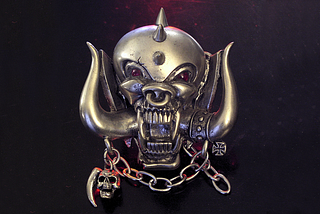 A belt buckle in the form of Snaggletooth, the symbol of the band Motorhead. It is a silver skull with large tusks and bared teeth with red eyes.