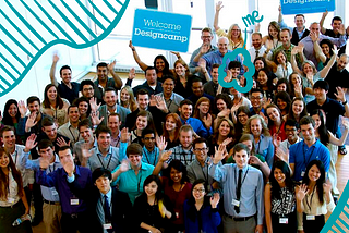A photograph of 70 people standing in an office and waving with lanyards on. A blue sign says “Welcome Designcamp.” A white woman with short brown hair is circled and notated with “me.”