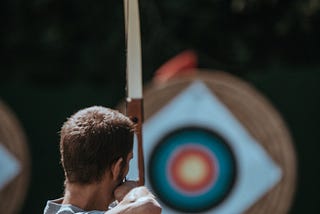 View of a man’s back as he uses a bow to draw an arrow, preparing to fire at a circular target in the distance.