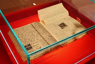 An open book with handwriting and a photo of Anne Frank sits in glass display case on a red platform.