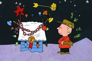 A frame from the cartoon “A Charlie Brown Christmas.” Charlie Brown, holding his thin, delicate little Christmas tree, looks with bemusement at Snoopy’s doghouse, which is decorated with gaudy lights and ornaments. Someone has placed a 1st Prize ribbon on the doghouse for a Christmas decoration contest.