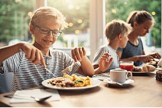 Ten year old boy sitting at the breakfast table in a modified chair with his brother and mother, happily eating with the family while also getting his sensory needs met in the chair that allows him to move.