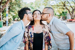 An adult woman standing in a sunny street, with two men either side of her each kissing her cheeks. Her face is split in a large grin, showing how happy she it with her situation.