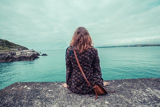 Young woman sitting on the edge of a cliff overlooking water.