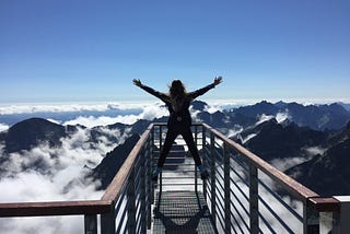 Person standing on hand rails with arms wide open facing the mountains and clouds