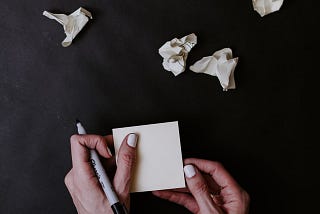 hands holding a permanent marker and sticky note pad with crumpled sticky notes above