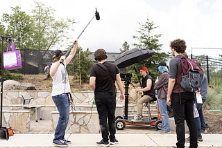 Several production assistants and an audio operator circle a camera operator working on a film/video project at an outdoors location