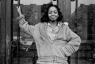 A young Black girl with brown skin and a proud smile stands in front of a Brooklyn brownstone in 1969.