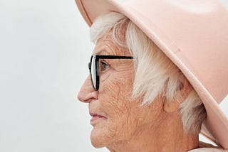 An old woman with short, white hair, a cream hat, and black-framed glasses sits in profile against a white background.