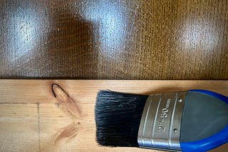 A paint brush beld against some bare skirting board. The background is a dark shiny wooden surface.