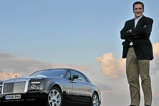 Imagen de Ian Cameron, diseñador de BMW y Rolls-Royce, con los brazos cruzados apoyado en el capó de un automóvil Rolls-Royce. El vehículo está estacionado con un cielo despejado al fondo durante lo que parece ser el atardecer o amanecer, dado el tono del cielo. La placa del vehículo es visible y muestra la inscripción ‘HX08 BHA’.