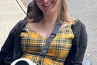 A young white woman with blonde hair and glasses, wearing a yellow plaid dress and purse with a skull, smiles outdoors in her power wheelchair.