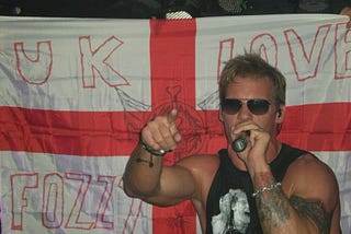 Chris Jericho of rock band, Fozzy, singing on stage in front of a St George’s Cross flag that says “UK Loves Fozzy”.