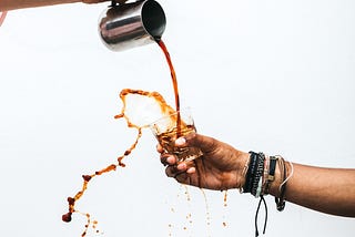 Coffee being poured from a metal jug into a small glass, splashing out over the top and over the hand of the outstretched arm holding the glass.