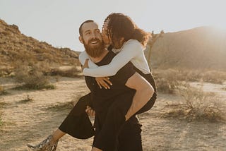 Man carrying his girlfriend on his back, as she kisses him on the cheek, in a desert sunset.