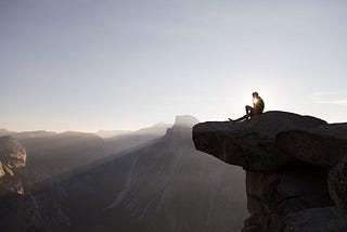 A person sits at the top of a cliff at sunrise