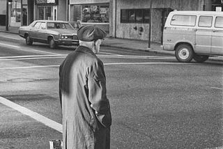 A Vancouver street scene, 1979, and elderly man in a cap and trench coat stands at a corner as cars pass.