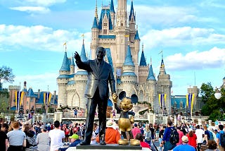A colorful photo of crowds at Magic Kingdom near the castle, with a statue featuring Walt Disney and Mickey Mouse in the foreground.
