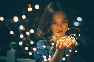 Asian woman gazes at a string of fairy lights in her hand; they cast a soft glow on her face.