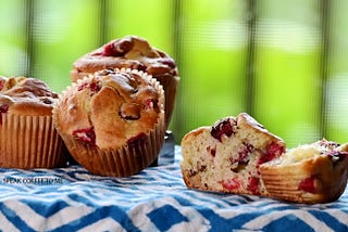 Four delicious sourdough cranberry nut muffins against a spring green background. One muffin in the foreground is cut in half and buttered.