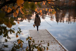 A person stands alone on a wooden pier overlooking a calm lake, surrounded by autumn leaves and tranquil reflections.