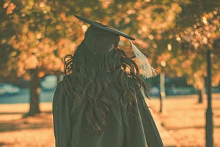 A woman wears a black graduation cap and gown with a gold tassel. She is walking away from the camera. In the background are several trees with fall leaves.