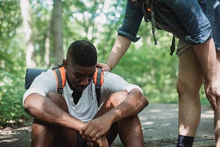 A young man is sitting on the ground with his head held down. Another man stands near him with his hand on the other’s shoulder.