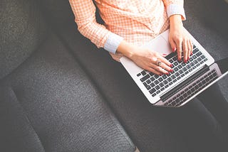 An overhead shot of a femme person in a checkered orange shirt. All that is visible is her white hands with red nailpolish and a ring typing on a Macbook. She is sitting on a grey couch.