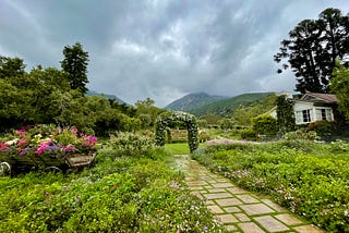 A scenic garden with a flower archway leading into the distant green mountains and semi cloudy blue skies.