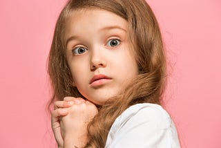 Child with long hair and big eyes. Their hands are folded under their chin, and they look worried or frightened.