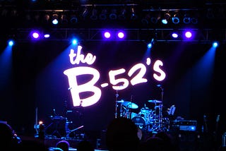 Stage with lit with spotlights and projection of B-52’s logo behind drum kit.