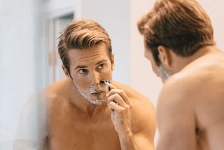 Man looking at himself in a mirror shaving. He has shaving cream on his face and is bringing a razor across his cheek.