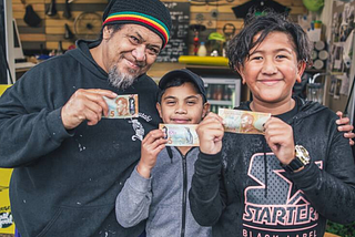 A man and two tama (boys) holding five dollar bills and smiling to camera