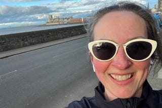 A sweaty woman with pink cheeks and white-framed sunglasses smiles at the camera. Behind her is a sea view and a road. She looks tired but happy.