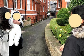 A woman is holding a baby. The baby is looking at an older child for the first time and they are all socially distanced. This is during COVID in 2020 in London. They are standing in front of a red-brick mansion,